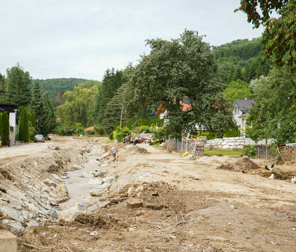 Eine vom Hochwasser stark beschädigte ländliche Straße mit einem Flusslauf, während Menschen vor Ort bei den Aufräumarbeiten helfen.