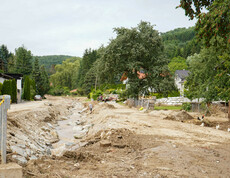 Eine vom Hochwasser stark beschädigte ländliche Straße mit einem Flusslauf, während Menschen vor Ort bei den Aufräumarbeiten helfen.