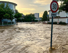 Eine überflutete Straße, auf der das Wasser bis zu den Verkehrsschildern reicht, mit einem Stoppschild im Vordergrund und Wohnhäusern im Hintergrund.