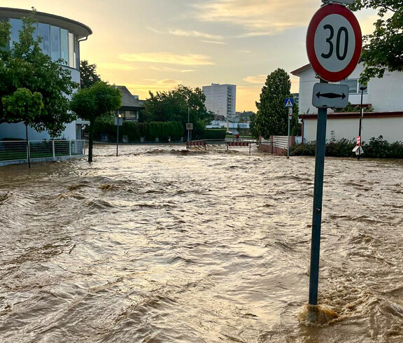 Eine überflutete Straße, auf der das Wasser bis zu den Verkehrsschildern reicht, mit einem Stoppschild im Vordergrund und Wohnhäusern im Hintergrund.