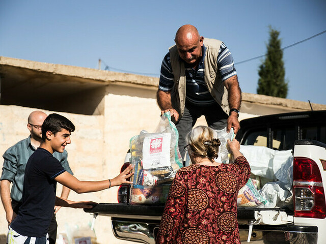 Ein Mann steht auf der Ladefläche eines kleinen LKWs und gibt Säcke mit Gewand an Menschen unten weiter. Auf den Säcken ist das Logo von Caritas Syrien.