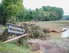 Hochwasserschäden im Südburgenland. Ein umgestürztes Schild und Äste liegen in einer überfluteten und schlammigen Landschaft.