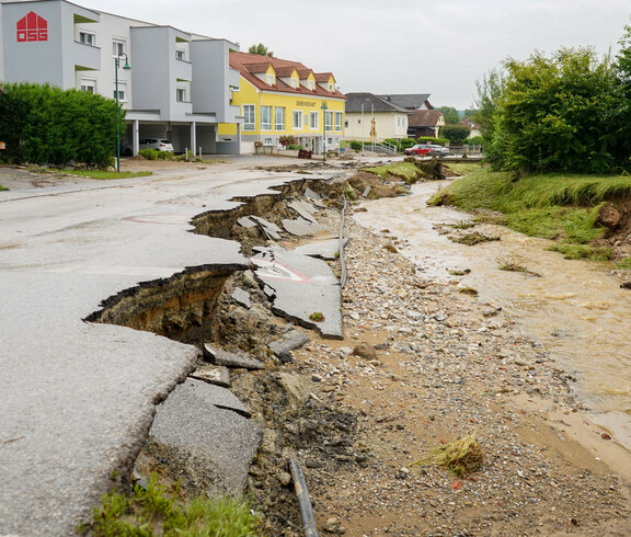 Eine von einem Hochwasser zerstörte Straße ist aufgebrochen, während ein überfluteter Bach neben der Straße fließt. Im Hintergrund sind Wohnhäuser zu sehen.