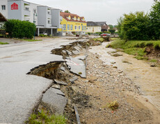 Eine von einem Hochwasser zerstörte Straße ist aufgebrochen, während ein überfluteter Bach neben der Straße fließt. Im Hintergrund sind Wohnhäuser zu sehen.