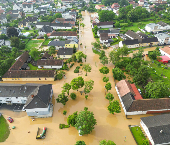 Eine Luftaufnahme zeigt eine überflutete Wohnsiedlung, in der braunes Hochwasser Straßen und Gärten bedeckt, während Bäume und Häuser aus dem Wasser ragen.