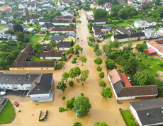 Eine Luftaufnahme zeigt eine überflutete Wohnsiedlung, in der braunes Hochwasser Straßen und Gärten bedeckt, während Bäume und Häuser aus dem Wasser ragen.