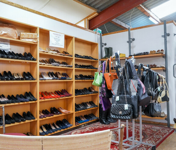 Ein Bereich im Second-Hand-Laden Carla Eisenstadt, in dem Regale mit einer großen Auswahl an Schuhen ordentlich präsentiert sind. Die Schuhe sind nach Farben und Typen sortiert, darunter schwarze, braune und rote Modelle. Neben den Schuhen hängen auf einer Kleiderstange Taschen und Kleidungsstücke, die ebenfalls farblich sortiert sind.