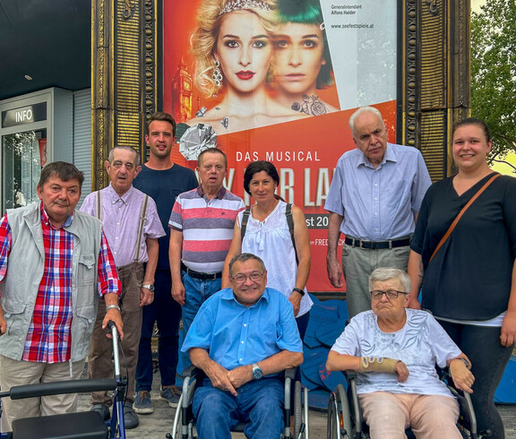 Bewohner*innen und Betreuer*innen des Haus St. Stephan waren zu Besuch bei dem Musical "My fair lady". Sie stehen gemeinsam vor den Musicalplakat