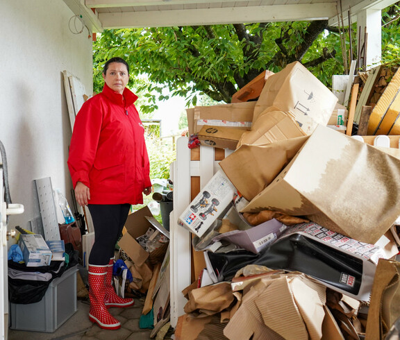 Melanie Balaskovics steht neben einem Stapel beschädigter Kartons und Habseligkeiten unter einem Vordach, umgeben von Aufräumarbeiten nach einem Hochwasser.