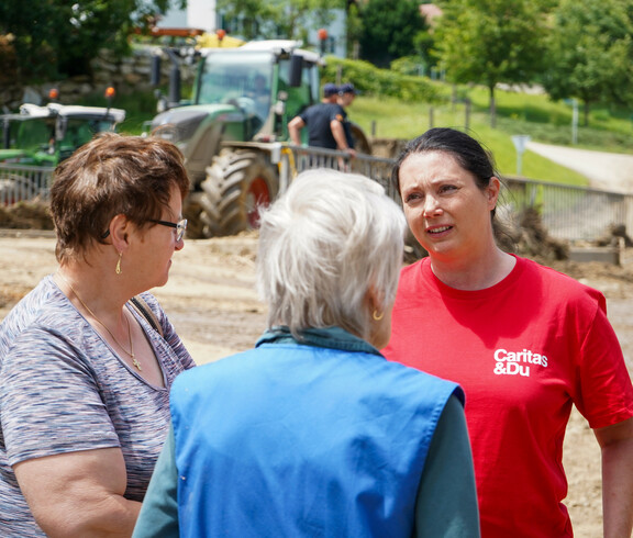 Melanie Balaskovics spricht draußen mit zwei vom Hochwasser betroffene Frauen, während im Hintergrund Bauarbeiten und Traktoren zu sehen sind.