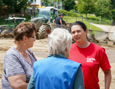 Melanie Balaskovics spricht draußen mit zwei vom Hochwasser betroffene Frauen, während im Hintergrund Bauarbeiten und Traktoren zu sehen sind.