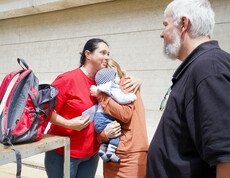 Melanie Balaskovics umarmt eine Frau mit einem Baby im Arm, während ein Mann daneben steht. Ein Rucksack liegt auf einem Holztisch im Vordergrund.