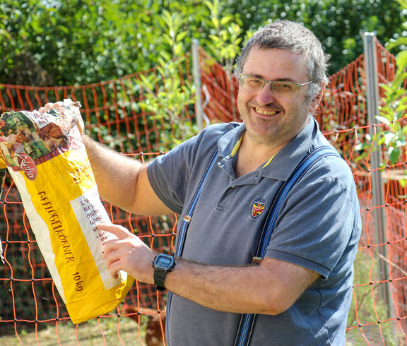 Ein Mann lächelt, während er eine Tüte Geflügelkörner hält. Er steht draußen vor einem orangefarbenen Zaun und füttert Hühner.