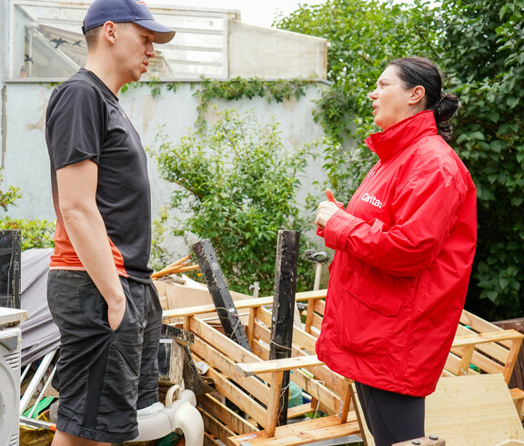 Melanie Balaskovics spricht mit einen von Hochwasser betroffenen Person. Dahinter sind Gegenstände zu sehen, welche durch das Hochwasser beschädigt worden sind.