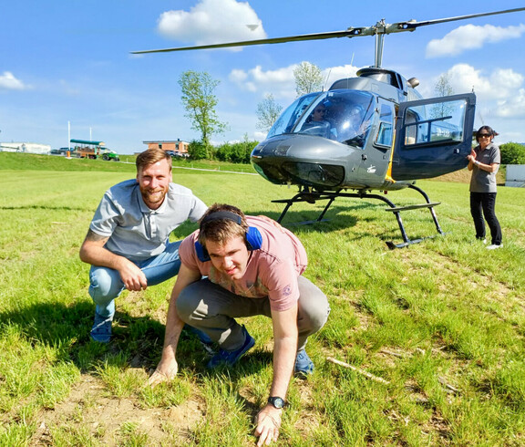 Ein Bewohner und Betreuer aus dem Haus St. Stephan knien auf einer grünen Wiese vor einem gelandeten Hubschrauber, während eine weitere Person neben der geöffneten Tür des Hubschraubers steht.