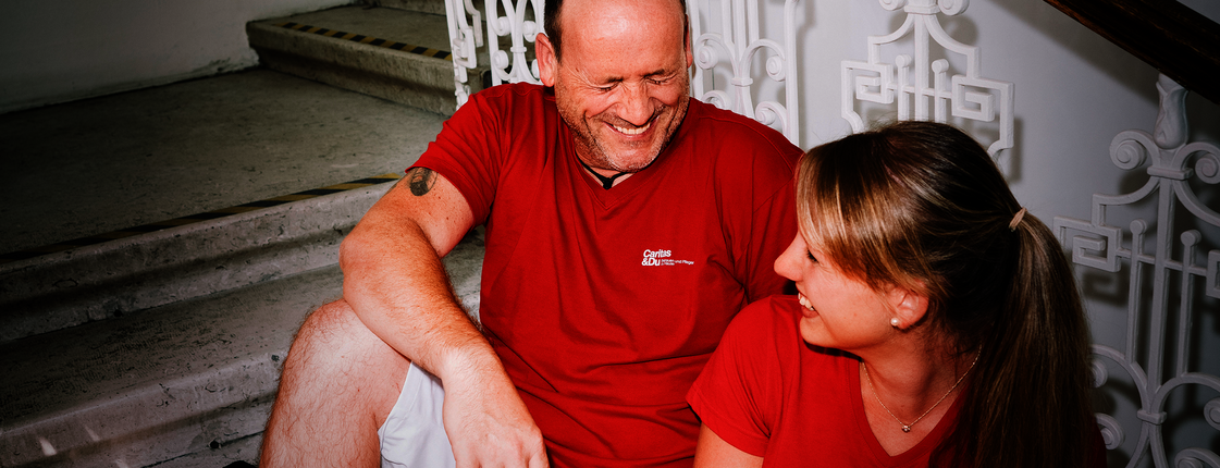 Ein Mann und eine Frau mit roten T-Shirts mit Caritas-Logo sitzen auf den Stiegen und lachen gemeinsam. Die Frau hat einen Aktenordner in der Hand.