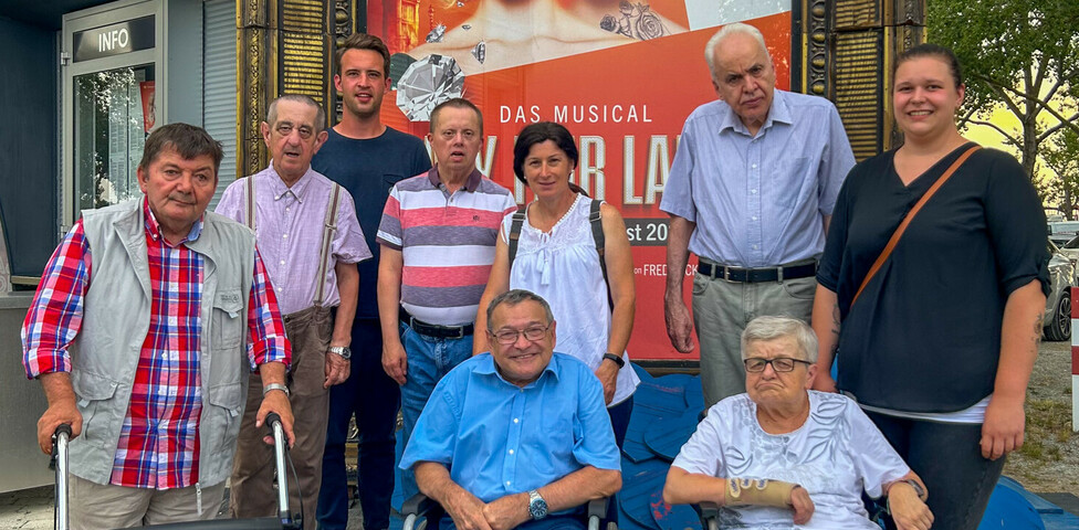 Bewohner*innen und Betreuer*innen des Haus St. Stephan waren zu Besuch bei dem Musical "My fair lady". Sie stehen gemeinsam vor den Musicalplakat