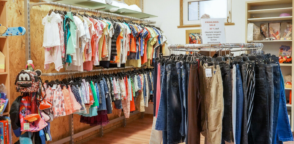 Second-Hand-Laden Carla Eisenstadt, wo verschiedene Kleidungsstücke, wie Hosen, Jacken und bunte Oberteile, auf Kleiderstangen ordentlich aufgehängt sind. Links im Bild befinden sich weitere Kinderkleidungsstücke und Accessoires. Im Hintergrund stehen Regale mit zusätzlichen Artikeln. 