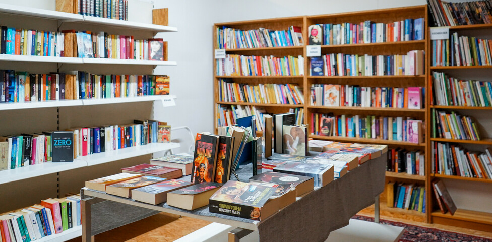 Der Bücherbereich der Carla Eisenstadt. Mehrere Regale sind mit einer breiten Auswahl an Büchern gefüllt, die ordentlich sortiert sind. Im Vordergrund befindet sich ein Tisch mit weiteren Artikeln, mit Accessoires oder Zeitschriften.