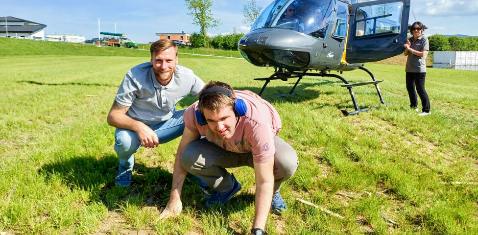 Ein Bewohner und Betreuer aus dem Haus St. Stephan knien auf einer grünen Wiese vor einem gelandeten Hubschrauber, während eine weitere Person neben der geöffneten Tür des Hubschraubers steht. Es herrscht sonniges Wetter.