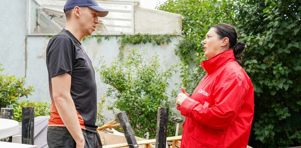 Melanie Balaskovics spricht mit einen von Hochwasser betroffenen Person. Dahinter sind Gegenstände zu sehen, welche durch das Hochwasser beschädigt worden sind.