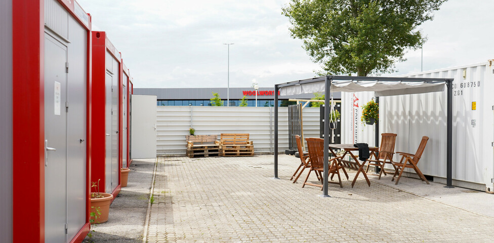 Mehrere rot-graue Container im Freien, gegenüber ein überdachter Sitzbereich.