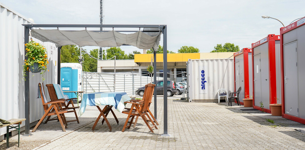 Bequeme Holzsessel und ein Holztisch mit Sonnenschutz. Im Hintergrund sieht man Container.